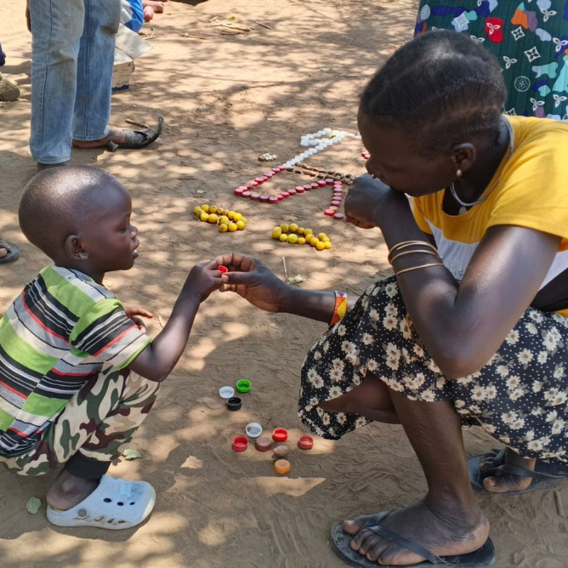 A teacher in Uganda sitting with a child passing them a bottle top