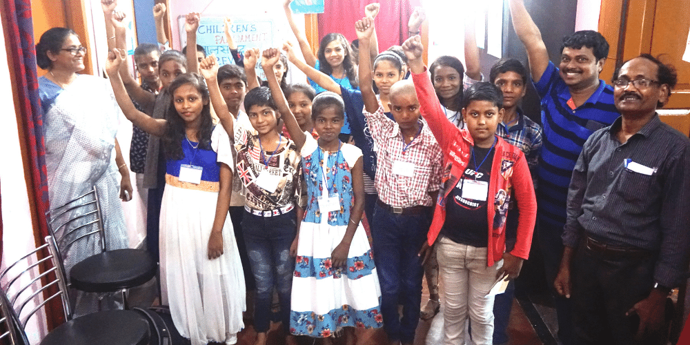 A room full of older Indian children holding their fists up to the air and looking at the camera. Some are smiling. Two men are standing on the right of the image, also smiling. A woman is standing on the left, also smiling towards the children. They all have lanyards round their necks with name tags and are members of the the child parliaments we support in India. 