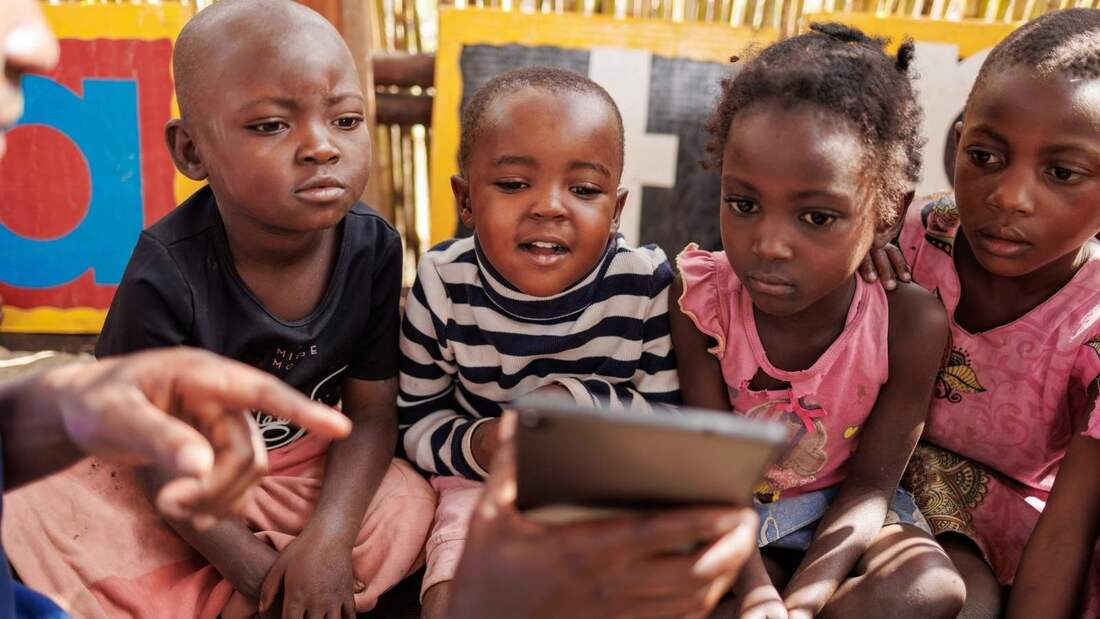 Four young Congolese children are sat looking at a tablet device being held in front of them in their shelter where they are taking part in a cluster learning lesson