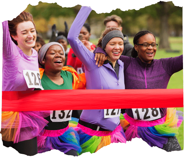 Four women dressed in purple wearing rainbow tutu's and running race numbers are crossing the finish line of a race together