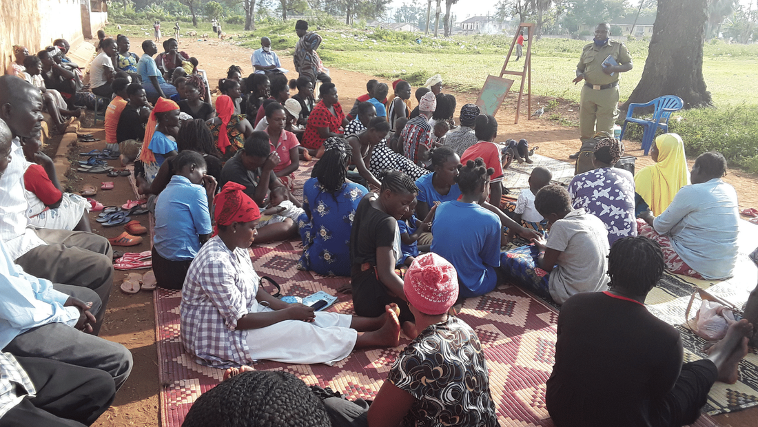 Community workshop taking place outside in loco slum community. Around 40 participants are sat on mats and benches and are listening to a police officer speak in front of them. 