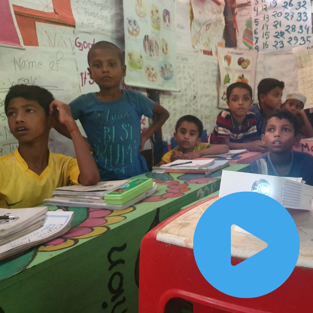Rohingya children sat in classroom watching video screen. Press to watch video