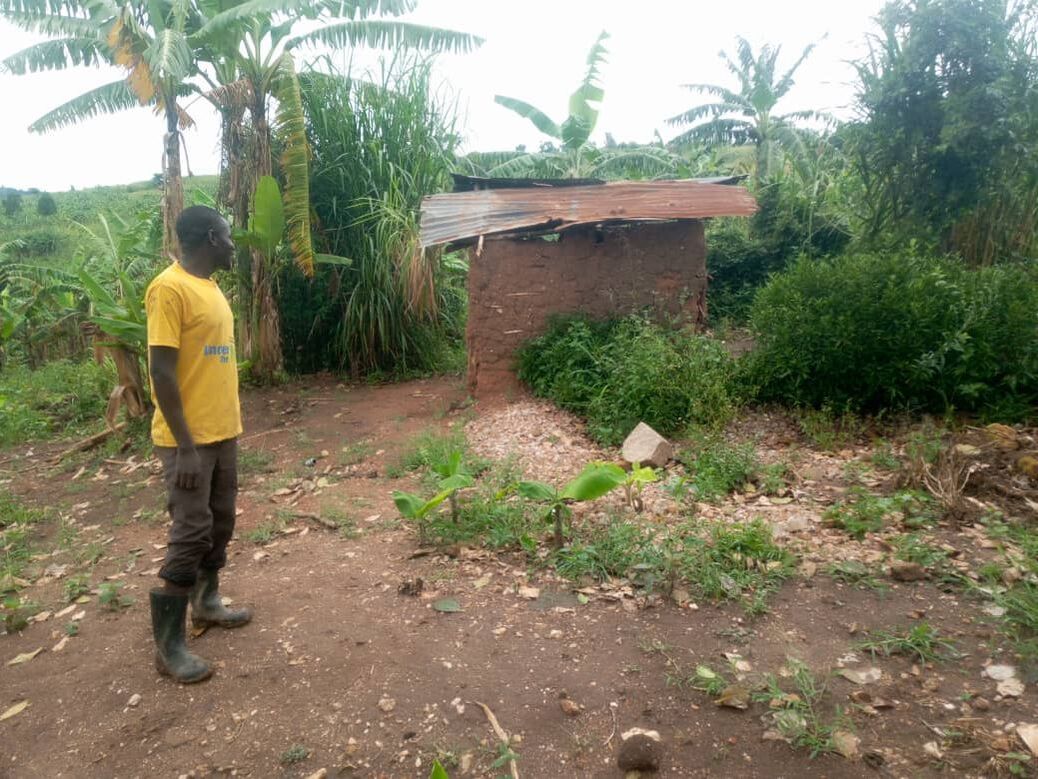 Joseph, a black man in his 20's wearing a yellow tshirt is standing looking at the dilapidated toilet hut at St Joseph's school. The toilets are in a small mud and wattle structure with a rusty corrugated metal roof 