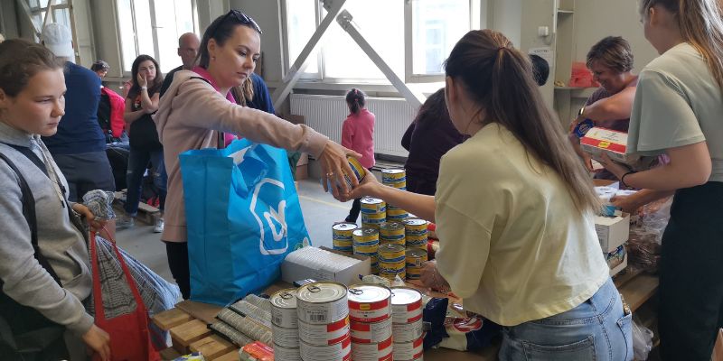 Two Ukrainian refugee women collecting tins of food from a volunteer table in a large white building