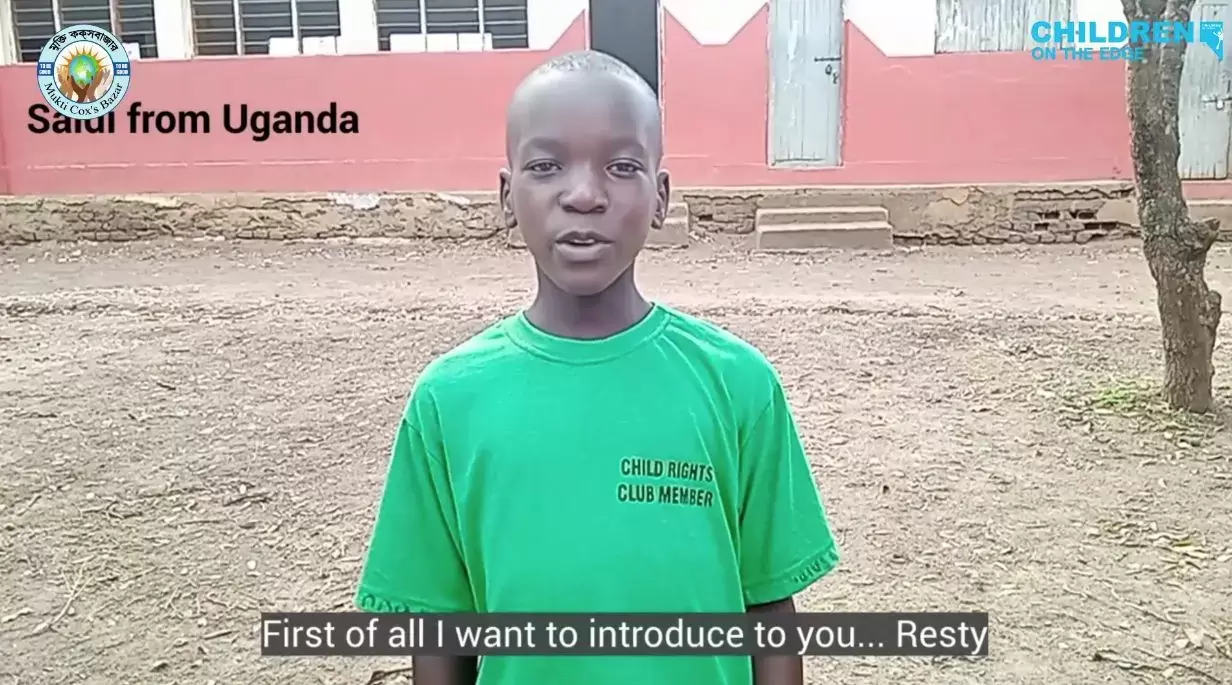 Boy from Uganda is talking to the camera against the backdrop of a red building