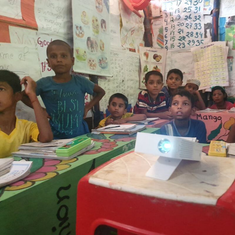 Children in a colourful classroom are watching a digital lesson behind a small white projector