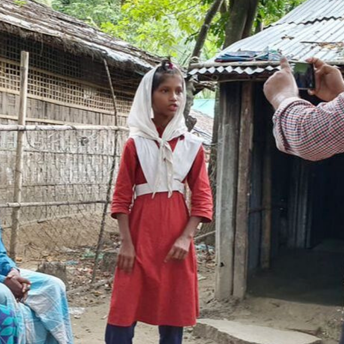A Bangladeshi girl being filmed talking outside her home as her parents sit beside her. 