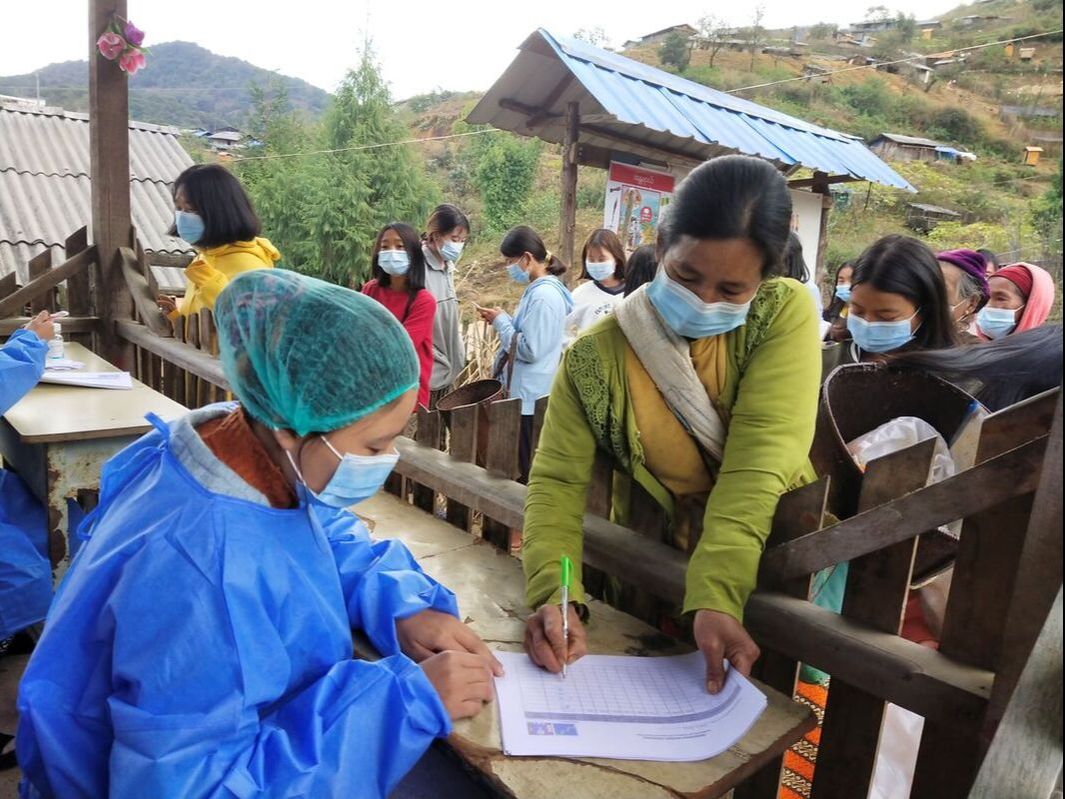 A Kachin woman is stood at the front of a queue outside and is signing a piece of paper being held by a woman in PPE at a wooden desk. 