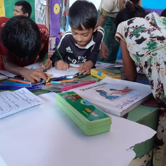 Three young Rohingya children in Bangladesh drawing and writing together on a table with colouring pencils