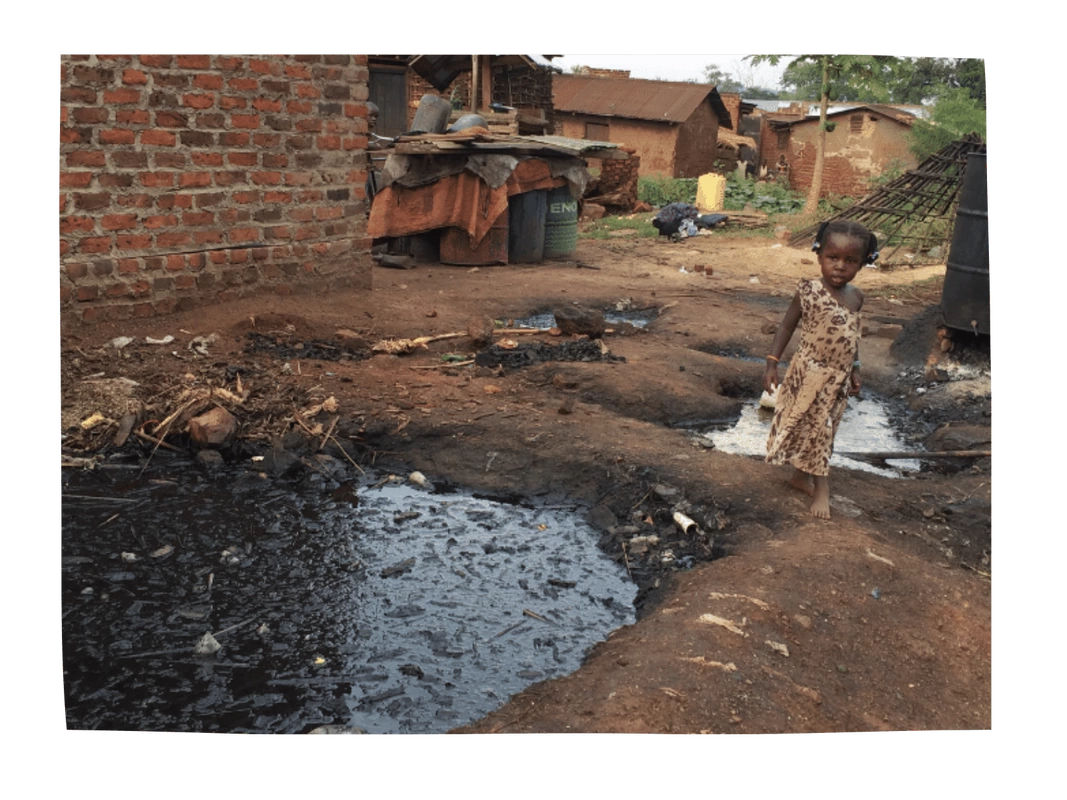 A young child is stood in a Ugandan slum next to a puddle of black sludge