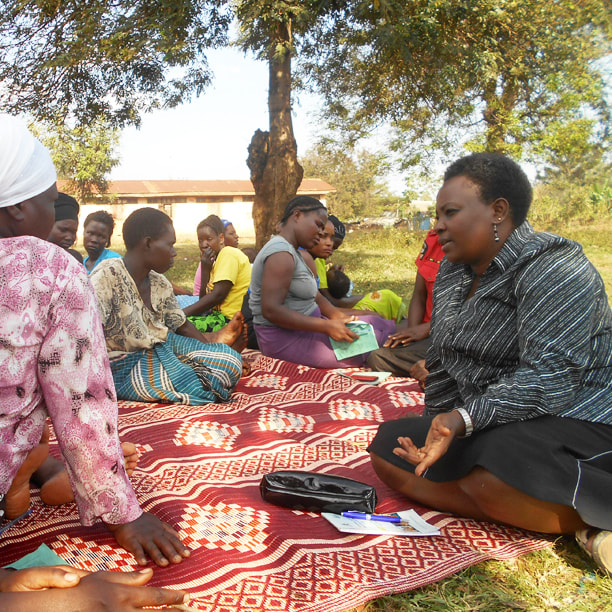Winnie, a black woman, sat on a colourful mat under a tree with a group of Ugandan women. they are in deep conversation.