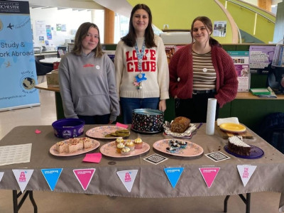 Three Students standing behind a table inside their university building selling cakes