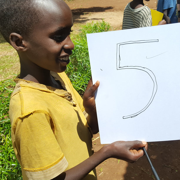 Ugandan boy wearing a yellow tshirt is holding up a written sign with the number '5' on it. Click on the image to read the  blog post.