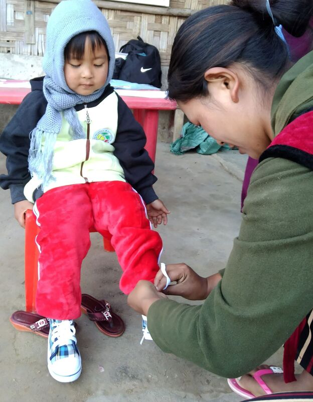 A young girl in Kachin is sat on a stool while an adult puts on a brand new pair of trainers.