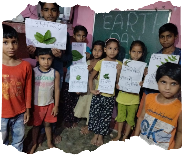 A group of younger Indian children and their teacher are holding up drawings they have made with green leaves in front of a blackboard that says 'Earth Day'.