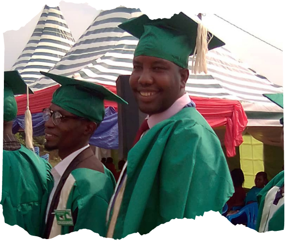 John, a black man wearing a green graduation gown and cap is smiling at the camera as he waits in line to collect his certificate. 