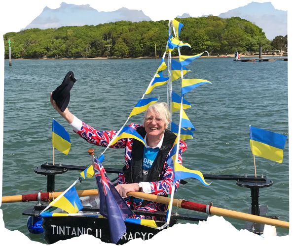 Major Mick, an elderly white man is sat in a tin boat covered with Ukrainian blue and yellow flags. He is on the water rowing and smiling, with his cap off in his hand.