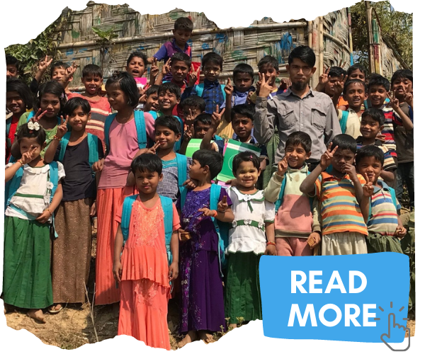 A group of Rohingya children standing together on a hillside outside their classroom in Kutupalong refugee camp. The children are wearing brightly coloured clothes and smiling and waving at the camera. You can click on the image to read more about working on 'the edge'.
