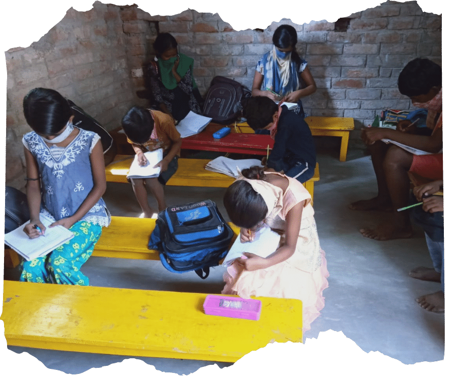 Small class of Indian students are sat doing work in a basic brick building. They are socially distance and all wearing masks. 