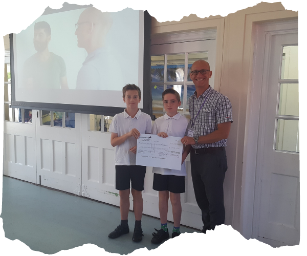 Children on the Edge COO, Ben is standing in a school classroom with two younger pupils holding a cheque and smiling