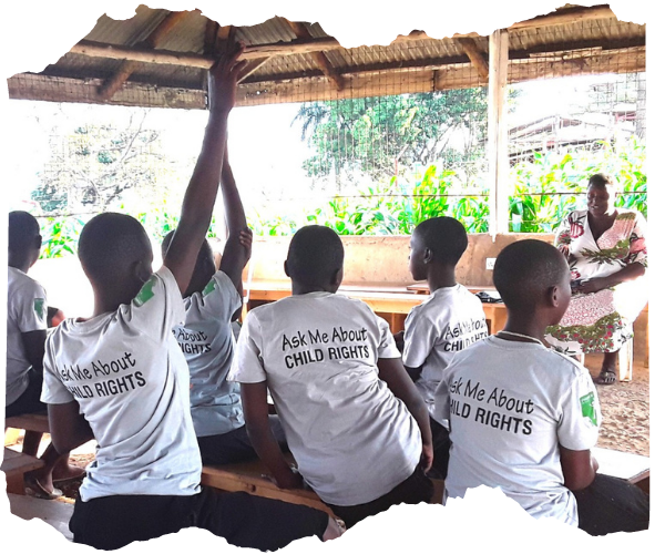 A group of Ugandan children are in a group listening to a speaker but putting their hands up to contribute. Their backs are to the camera and they are wearing white T shirts that say on the back 'Ask me about child rights'. 