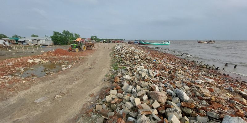A tractor is on a dirt road on the shore of Bhasan Char Island. There are bricks and rubble forming sea defences along the shore line with traditional boats moored up in the distance. 