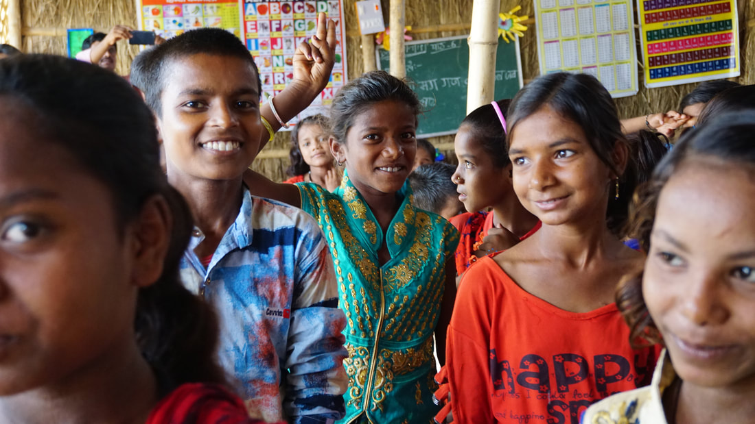 Two Indian girls standing at the front of their class facing the camera. They are both smiling. The girl on the left of the image is wearing a blue and gold dress with a red and white scarf. The girl on the right has a white and blue striped top with blue dungarees worn over the top. They are standing in front of a room full of other students who are sat down, most are looking in another direction. There are posters on the wall of the classroom showing numbers and letters. 