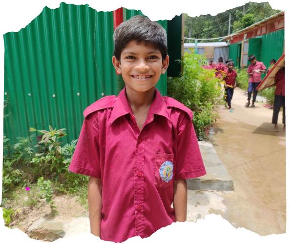 Borhan, a young Bangladeshi boy is wearing a red school shirt outside a green corrugated school classroom. He is smiling