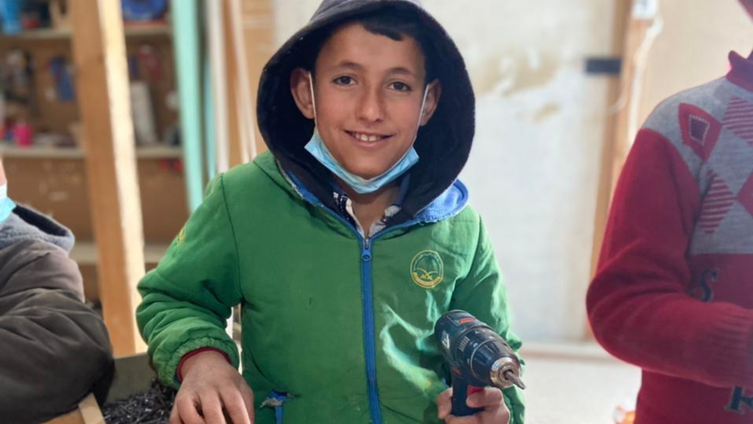 A Syrian boy is wearing a green zip up hoody and holding an electric drill in a carpentry class in Lebanon