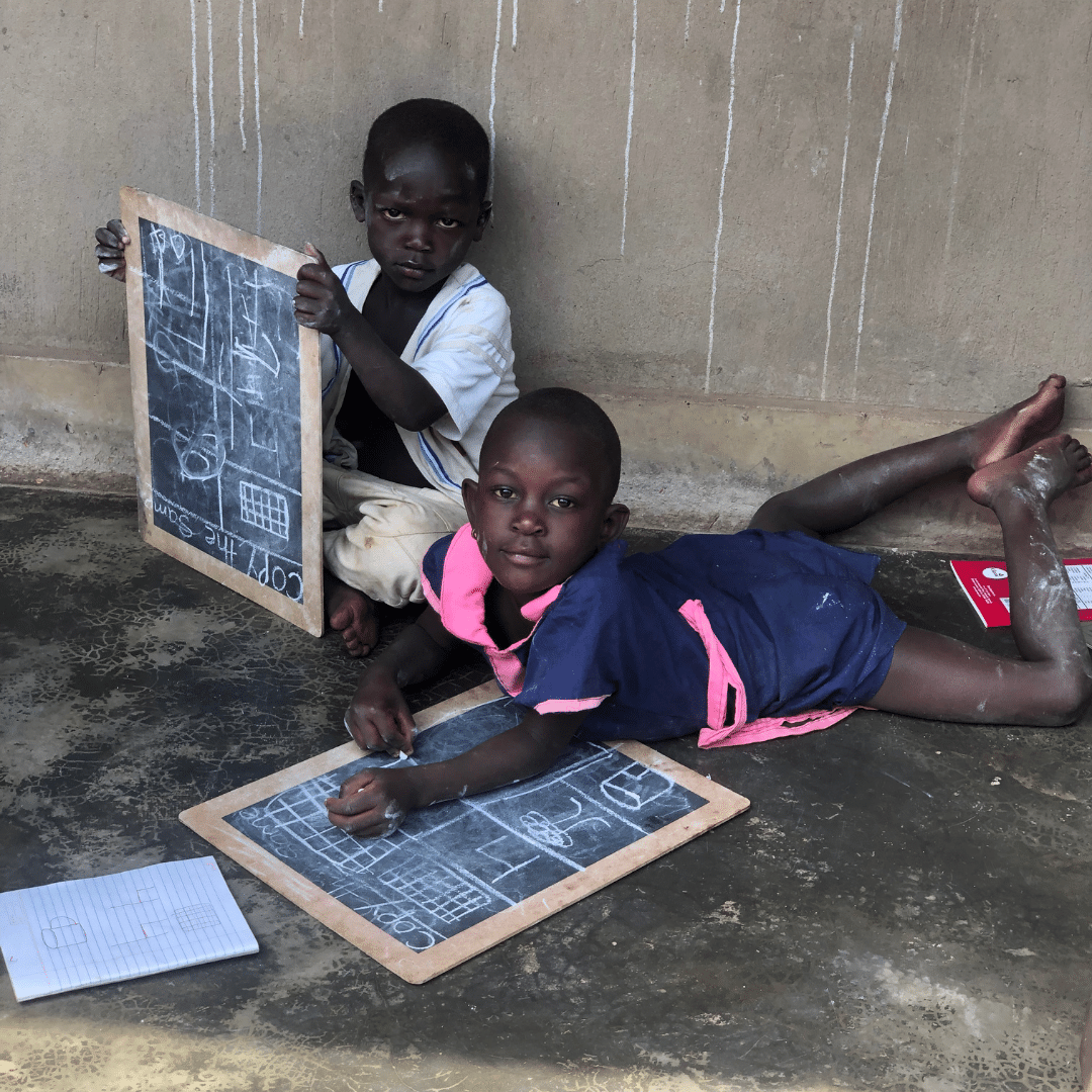 Two young Ugandan children writing on slates with chalk outside their home