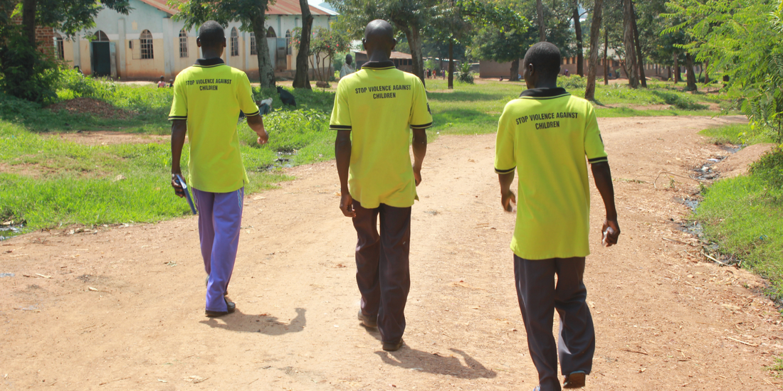 Three Ugandan men, who are members of a Child Protection Team. They are walking along a dirt track towards a large white building that looks like a church. They are walking away from the camera, wearing bright yellow tshirts which say: 'Stop Violence against children' on the back. Ahead of them is an area of grass with some tall trees.