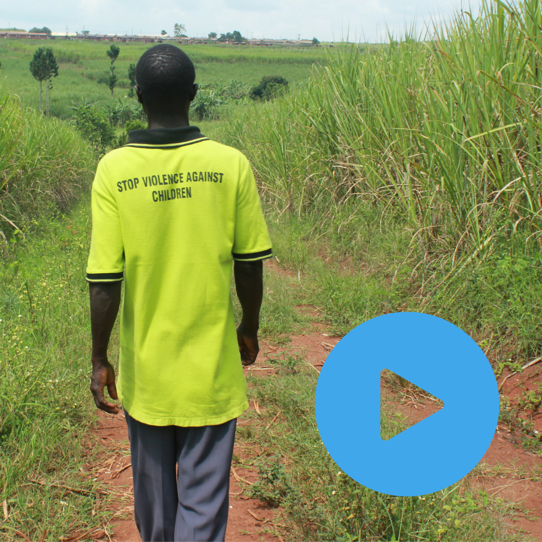 A Uganda man in a bright fluorescent T shirt is walking away from the camera down a grassy path. The back of his T-shirt says 'End violence against children'. This man is a member of one of the Child Protection Teams Children on the Edge support in Uganda. The image has a 'video play' icon as you can click the image to be taken to a video on Youtube about the Child Protection Teams. 