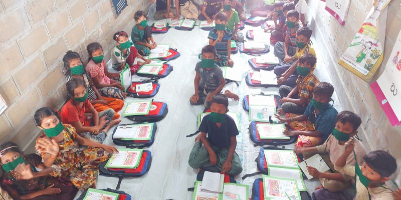 A brick classroom with while floor is full of Rohingya students, all wearing matching green facemasks. They are sat on the floor with their bags and school books open in front of them. 