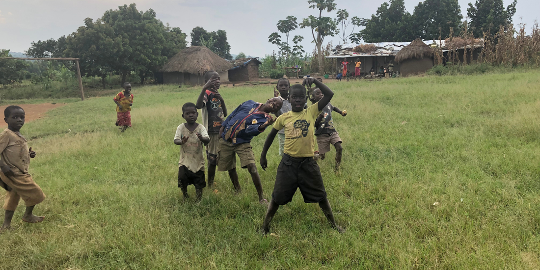 8 young Congolese boys playing in grassy space. There are crowded together and smiling at the camera. Two boys are slightly further away and running towards the group of boys. In the background are some basic mud huts with straw roofs. Two adults are standing in front of one of the larger buildings. Trees surround the houses in the distance.
