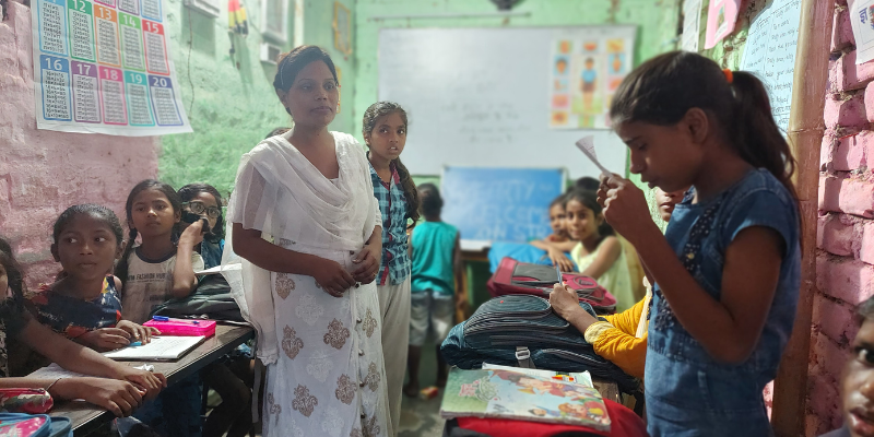 A Dalit girl wearing a denim dress is reading aloud in her classroom among her classmates and teacher who is looking at her