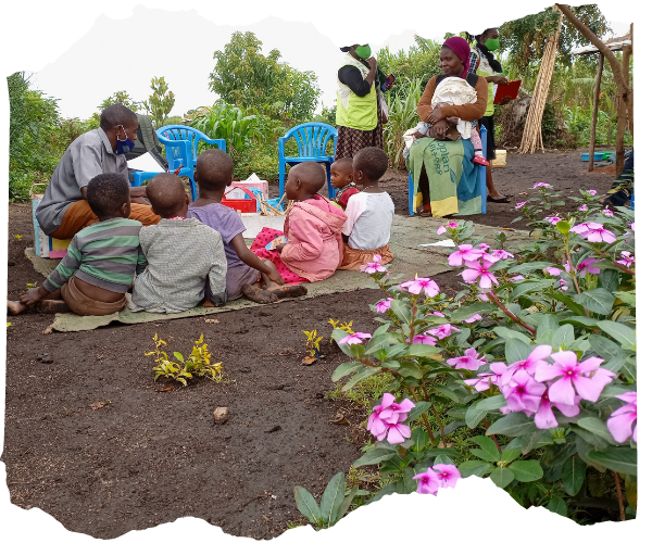 Group of Conglese children sit outdoors on ao mat with a male teacher having an early learning lesson. A Mother is sat with a smaller baby on a chair next to them, and a few Children on the Edge Africa staff are in the background, In the foreground is a bush of pink flowers and behind them are trees. 