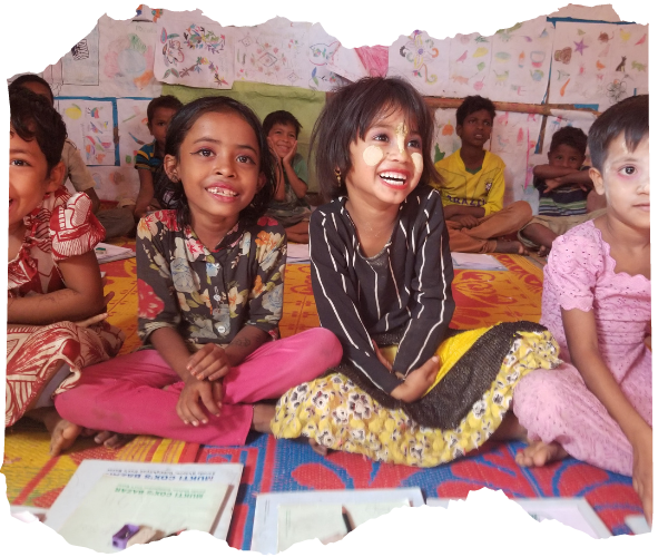 A line of Rohingya girls in a classroom packed with artwork on the walls. They are laughing and smiling and have their classwork on the floor in front of them. 