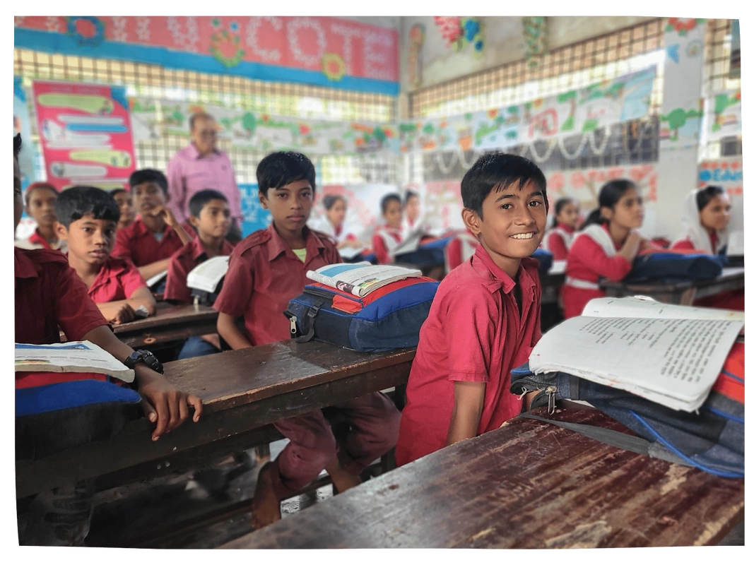 Bangladeshi classmates in red and white uniforms with projector, watching a video lesson and smiling. There is a button to clid to go back to the 'How we help' page.
