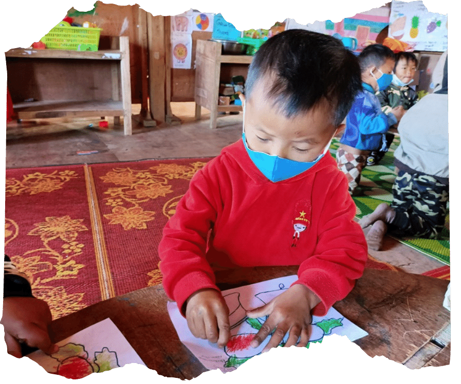 Young Kachin boy wearing a red jumper is sat in his classroom doing some colouring. He is wearing a blue facemask.