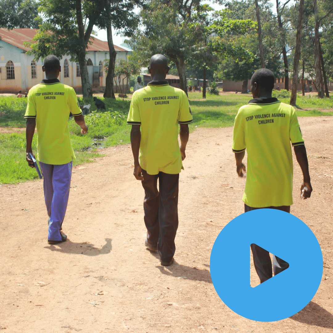 Three Child Protection Team members in Uganda are walking away from the camera down a dirt road towards some trees. On the back of their tshirts is written 'stop violence against children'. You can click on the image to watch a video. 