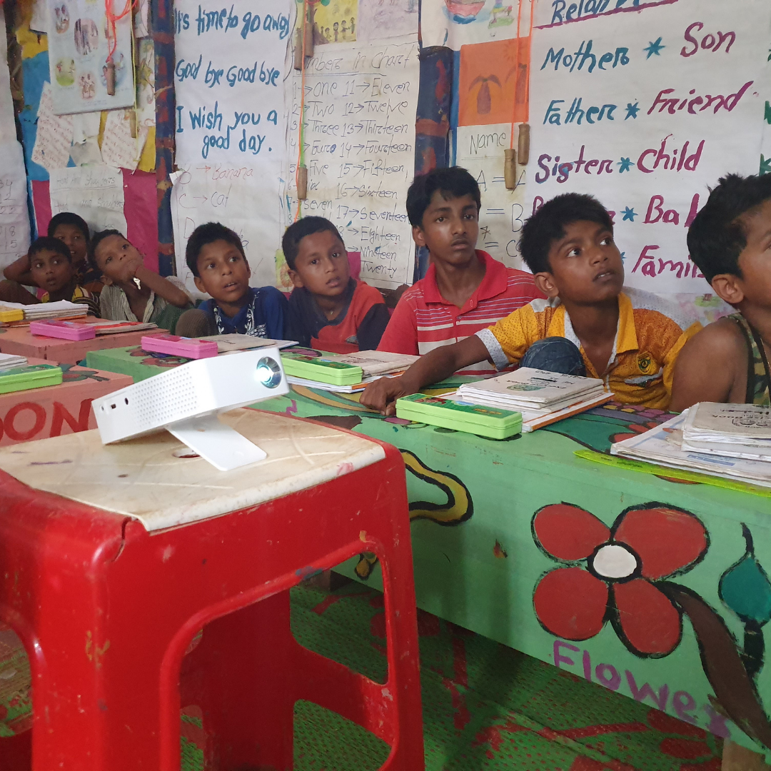 Image shows a line of young Rohingya refugee boys looking up at a video projection lesson. You can't see the projection, just their faces, and they are all captivated. The small white projector is just in front of them. You can click this image to read a story about the digital education programme in Kutupalong winning an award. 