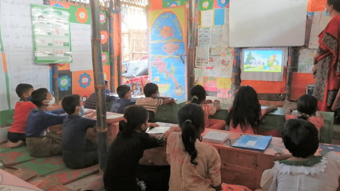 Rohingya refugee children in their classroom in Kutupalong camp are sat at their desks looking at a small projected cartoon at the front of class