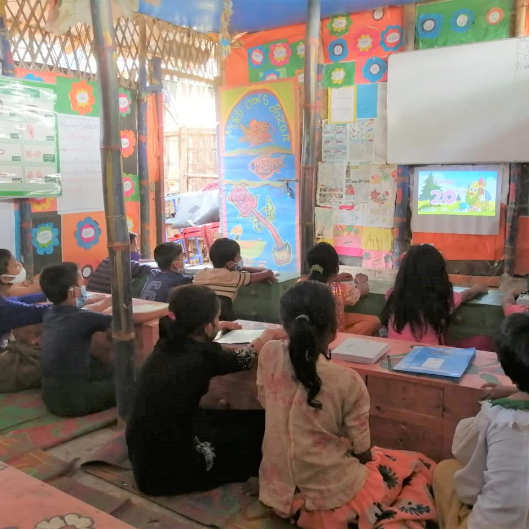 Digital lessons taking place in a classroom in Kutupalong. Children are sat at their desks looking at a projector screen.