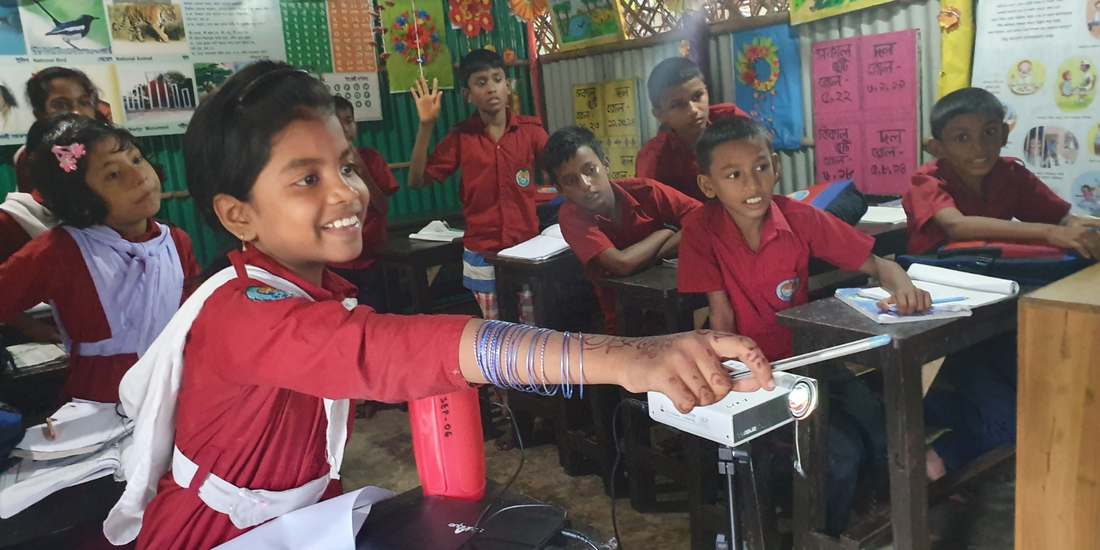 A classroom of happy Bangladeshi students sat at their desks, a digital projector is showing a lesson on the screen in front of them. 
