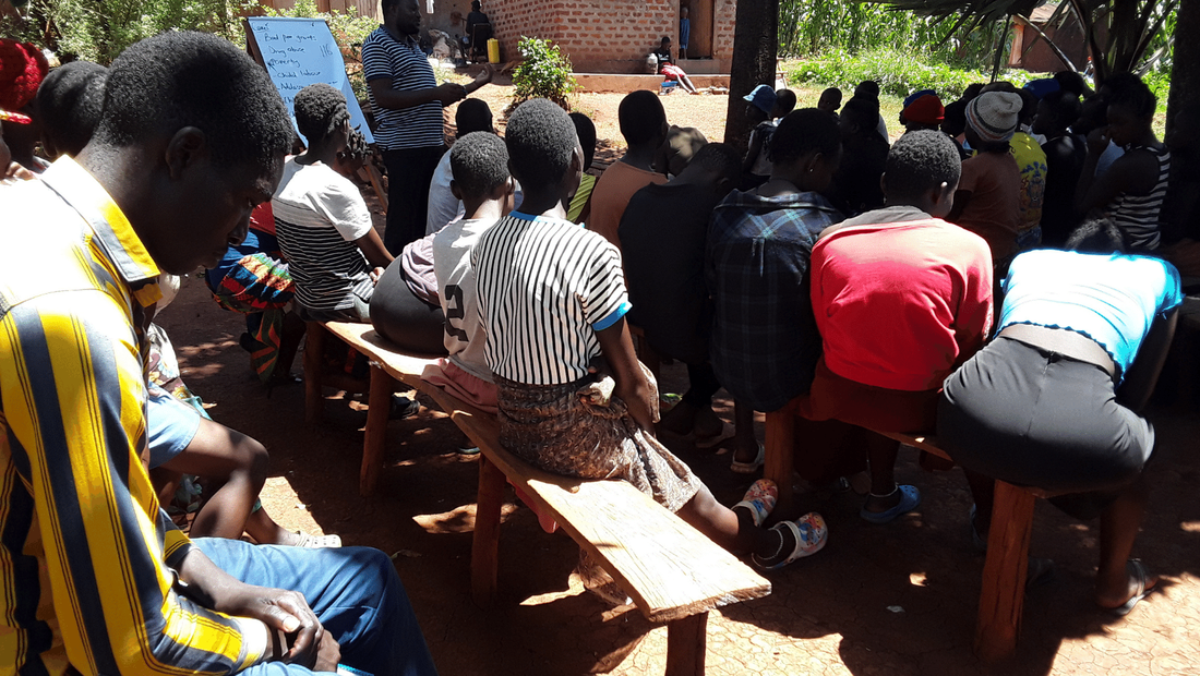 A community workshop taking place outside under trees in Masese III. Parents are sat on wooden benches listening to a CPT member lead the workshop.