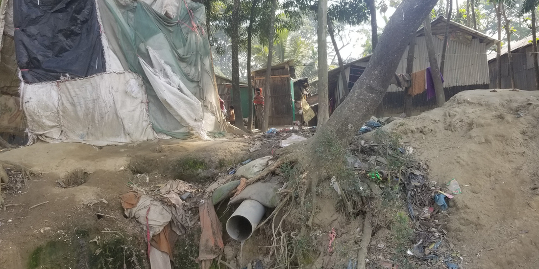 Makeshift shelters of tarpaulin and corrugated iron on the edge of a dusty riverbank covered in litter. 