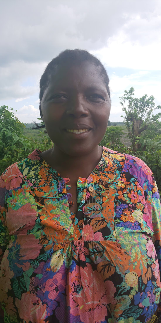 Furaha Dusabe, a middle aged black woman looks at the camera, smiling and wearing a bright floral shirt. Trees and green fields are visible in the background. 