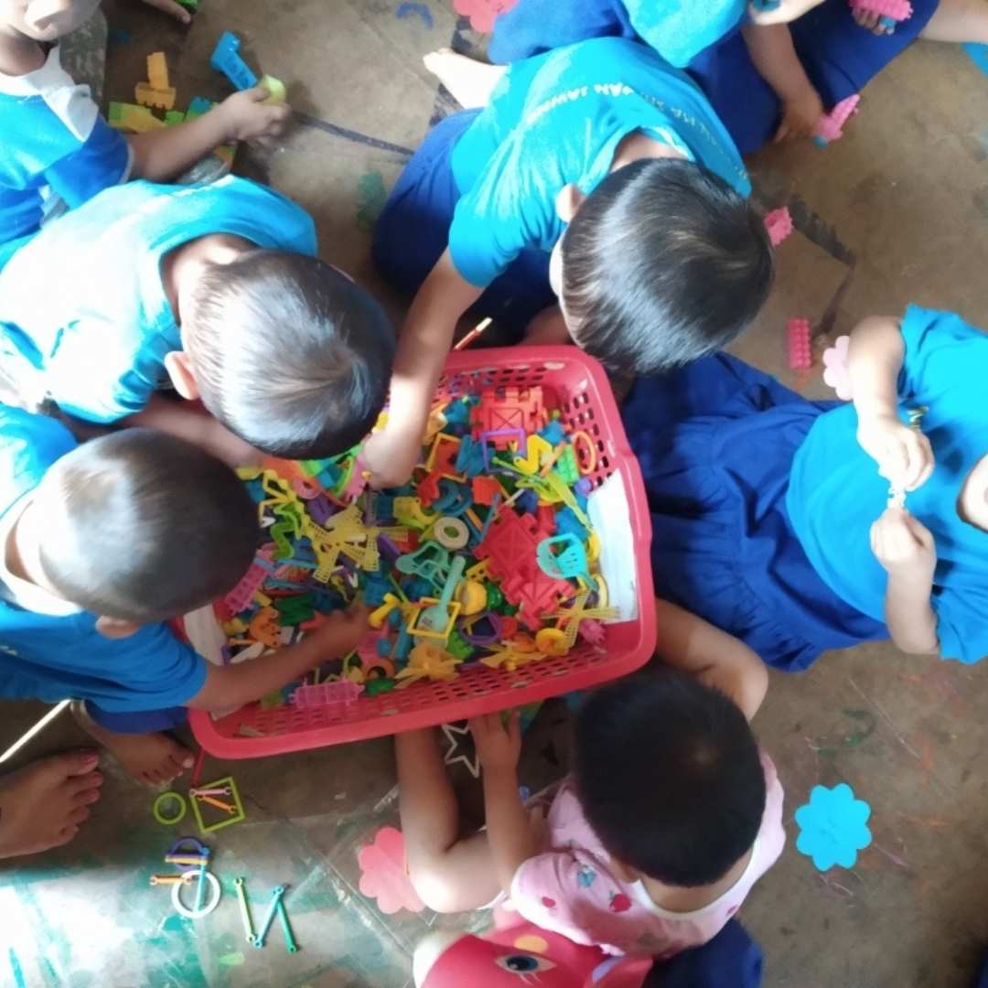 Group of young children from Kachin State, Myanmar,  taken from above, showing them surrounding a basket of colourful plastic shapes and choosing ones to take out and play with. 