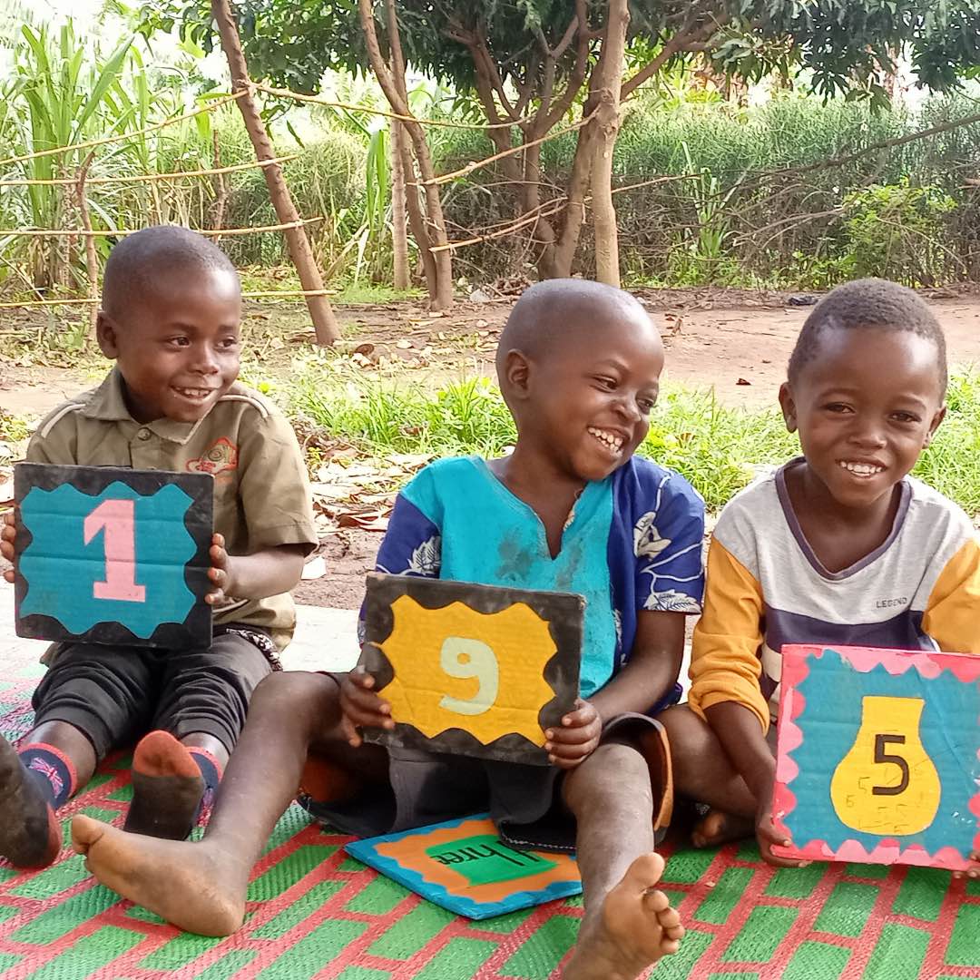 Three young Congolese boys sat in a line laughing and holding colourful signs with different numbers on, which are learning aids for their classes. 