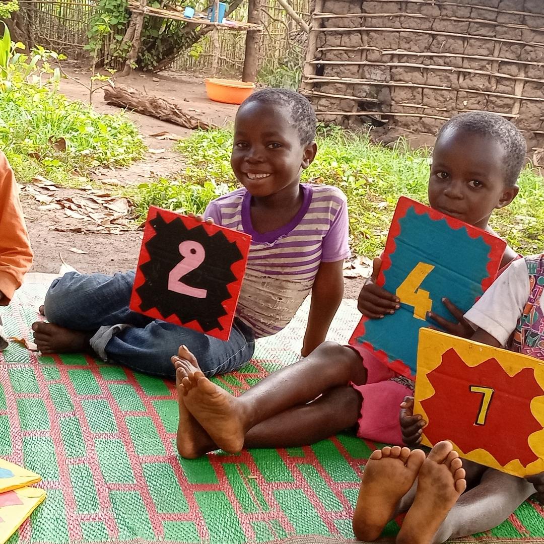 Three Congolese boys of around 5 years old are sat learning outside. They are holding up painted cardboard signs with numbers on them. 